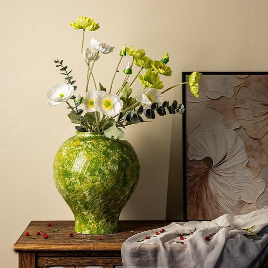 Green textured vase with flowers on a wooden table against a beige wall with a floral painting.