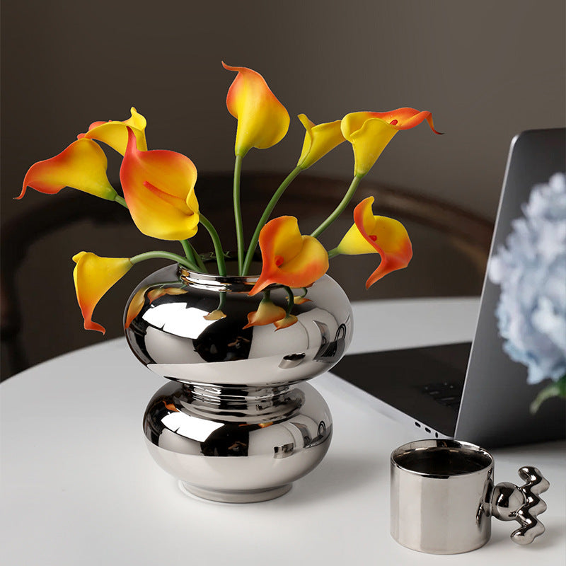 Double Orb Vase, reflective chrome-finish ceramic with rounded stacked form holding vibrant yellow and orange calla lilies