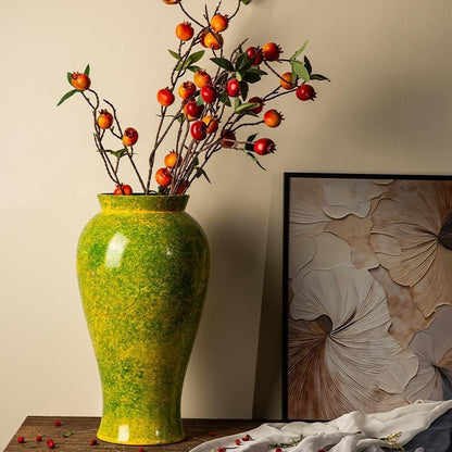 Green vase with decorative branches on a table against a wall with floral art.