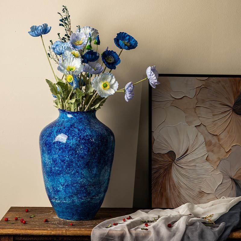 Blue vase with flowers on a wooden surface next to a decorative pillow against a beige wall.